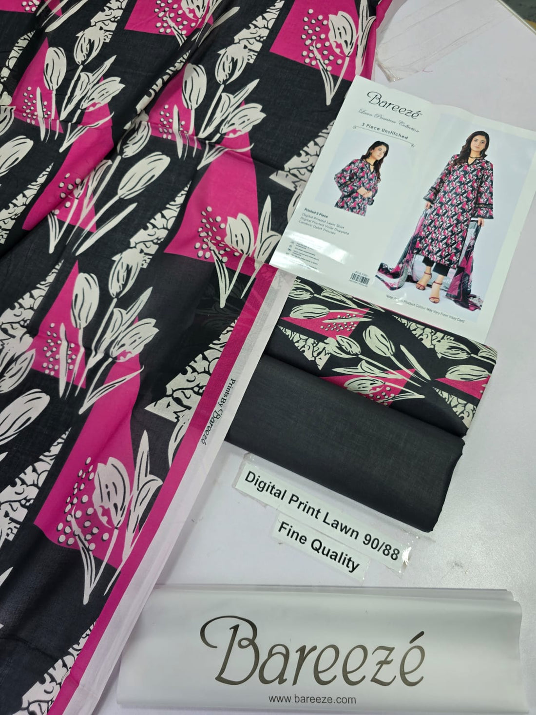 A model wearing a Bareezé 3-piece unstitched lawn collection set, featuring a digital printed lawn shirt, digital printed voile dupatta, and cambric dyed trouser. The set has a black base with pink and green floral patterns.