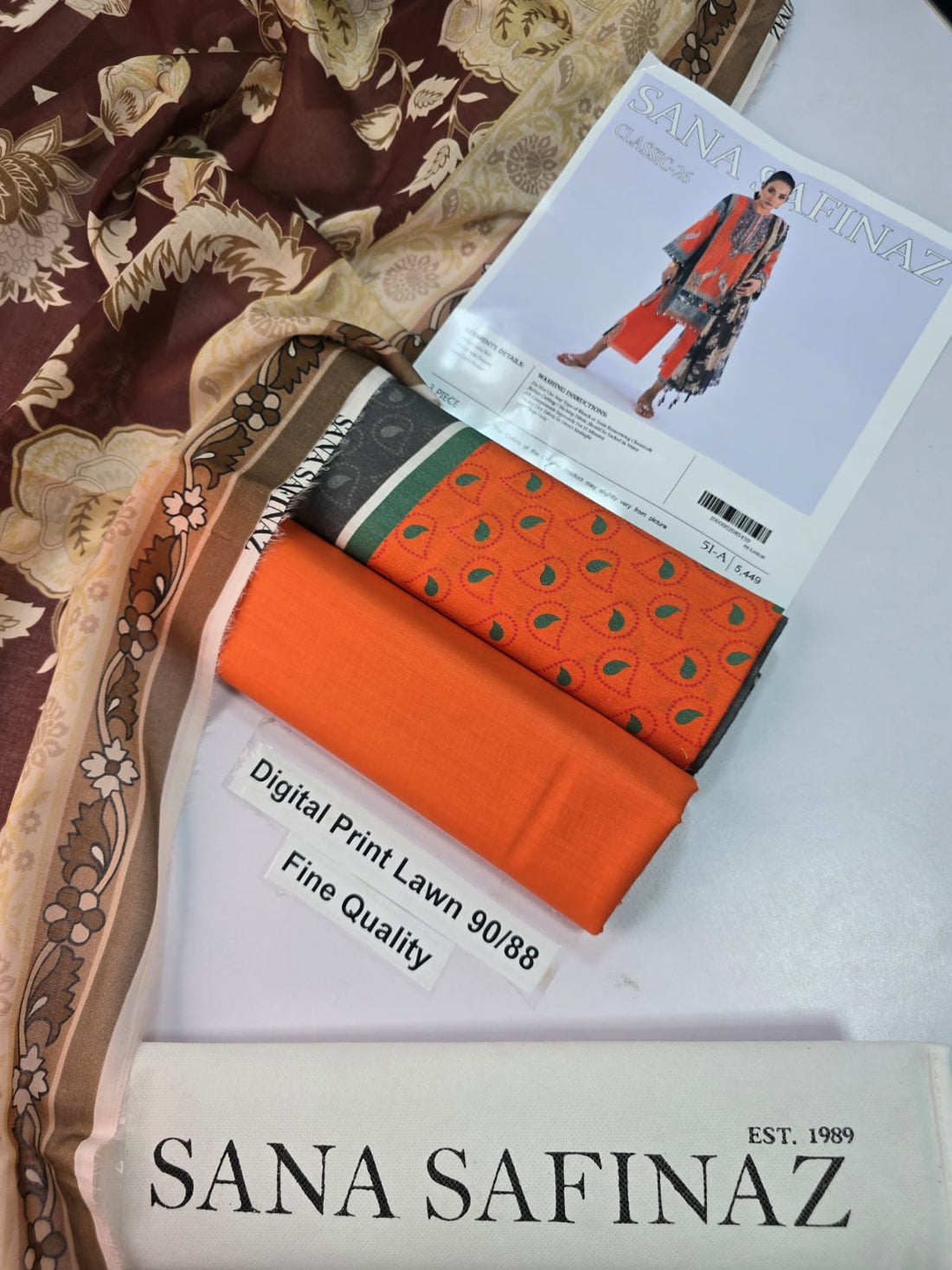 Sana Safinaz Classic-26 three-piece women's suit set featuring a printed kurta in orange, black, and beige with a matching dupatta, shown on a model against a light lavender background with product details at the bottom.